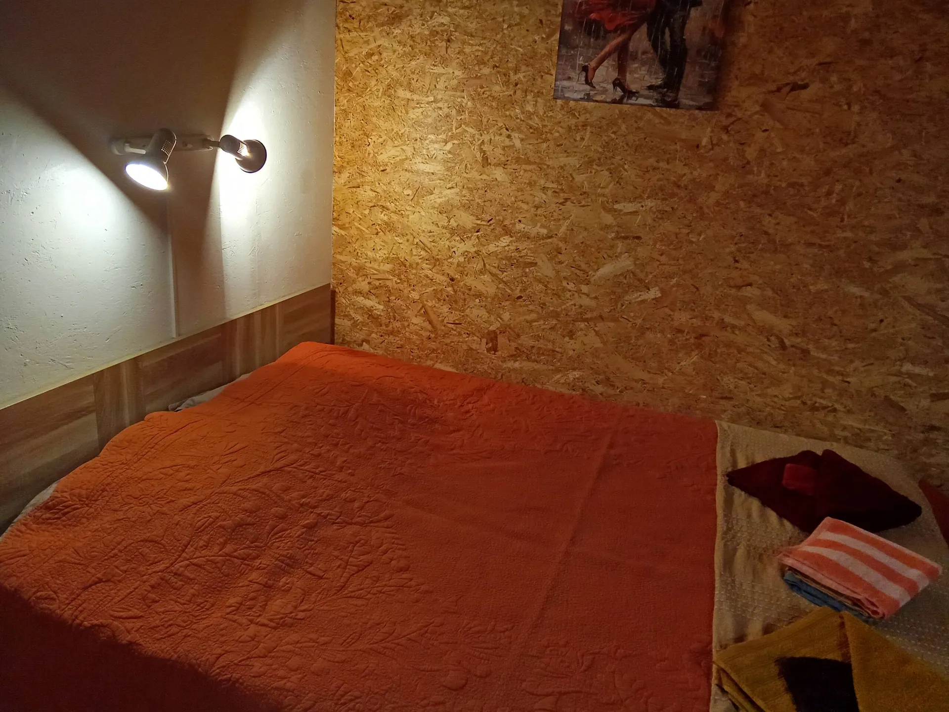 La chambre : lit double au couvre-lit orange, applique murale et paroi de bois