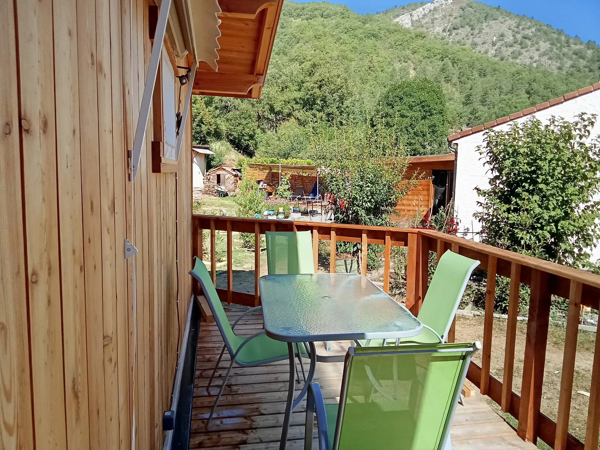 La terrasse en bois, sa table en verre et ses chaises vertes face à la montagne